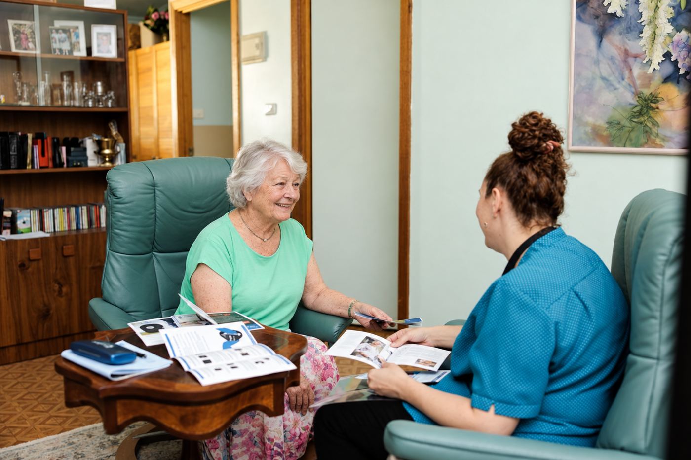 A resident and a carer sitting together in armchairs, looking through books and magazines.