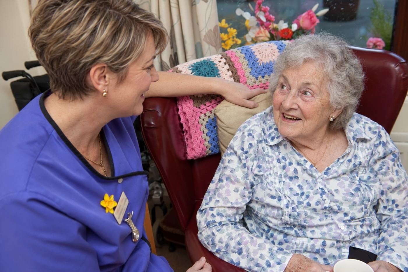 A carer kneeling beside a resident in an armchair, the two of them smiling at each other.